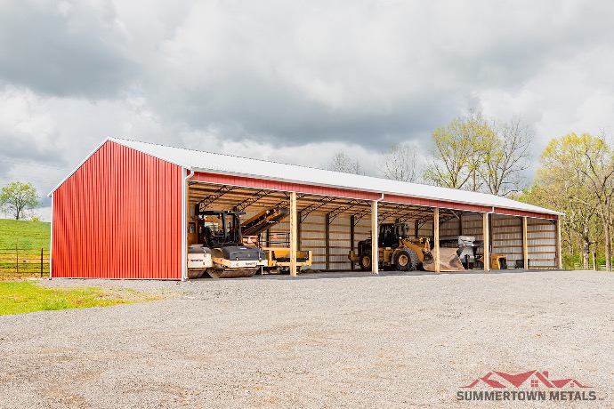 Three-sided hay barn with red siding, white roof, and heavy farm machinery stored underneath