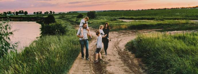 Happy rural family with three children walking down dirt road near lake and green meadow